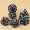 A collection of seven polyhedral dice in black and white are arranged on a light beige surface.
