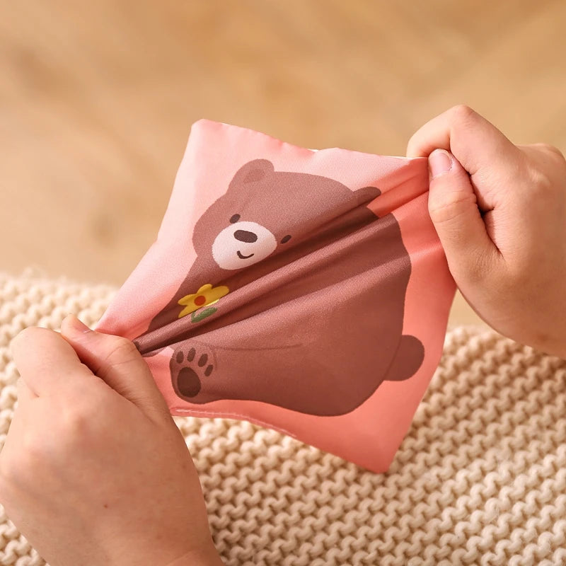 A hand is holding a soft fabric tissue box sensory toy shaped like a brown bear with a yellow flower on a beige knitted surfa