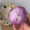 A translucent purple stress ball filled with glitter and small candies is held in a hand against a light background.