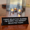 A black desk sign with white text humorously stating I have selective hearing and you weren't selected today.