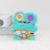 A bright turquoise monster plush toy with purple and orange eyes and an orange belly, sitting on a white surface.