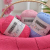Three skeins of pink, lavender, and blue cotton knitting yarn are displayed in a woven basket.