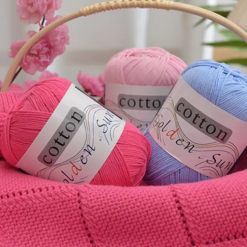 Three skeins of pink, lavender, and blue cotton knitting yarn are displayed in a woven basket.