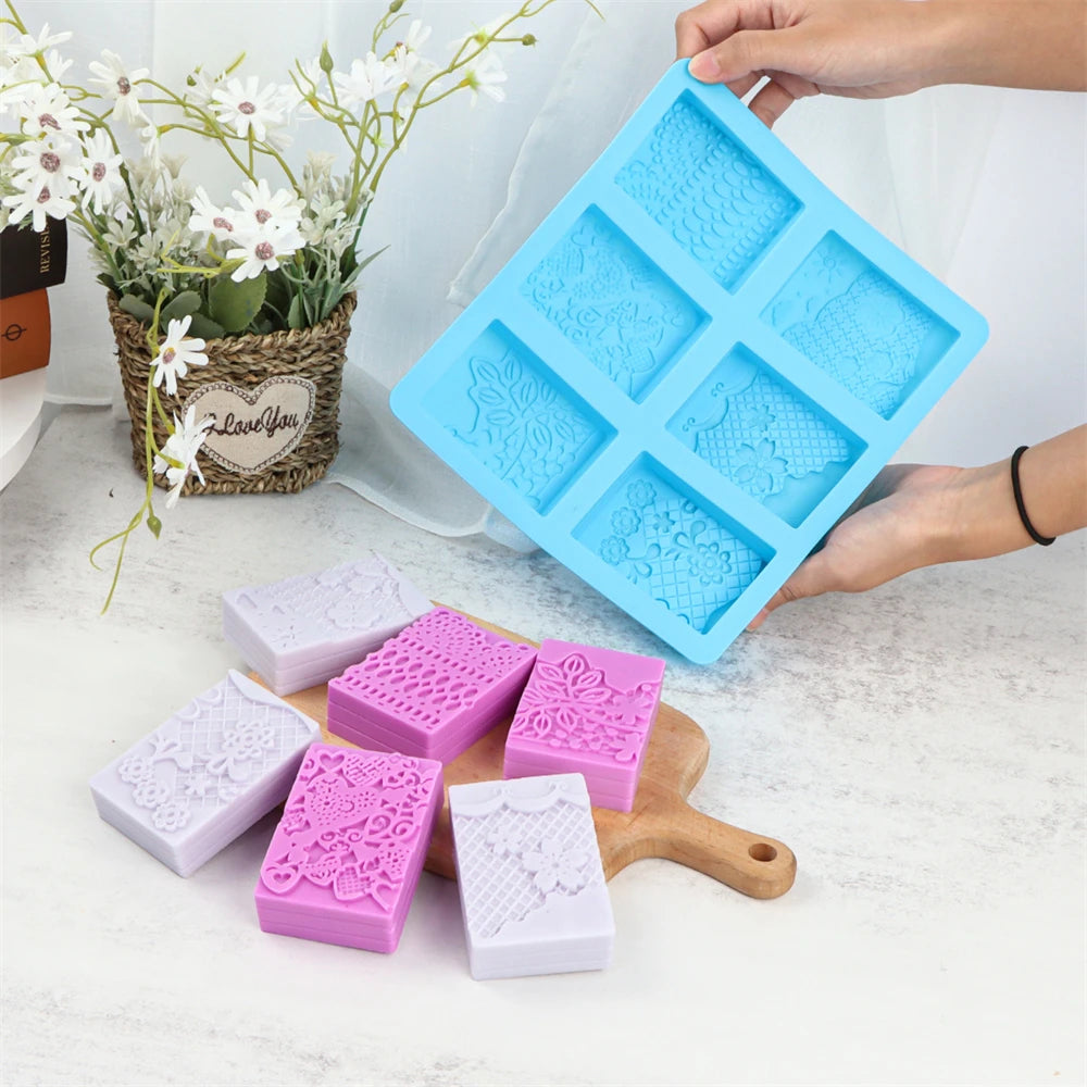 A blue silicone soap mold with six lace patterns is held by a hand, alongside several pink and white handmade soaps.