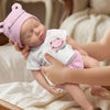 A realistic reborn baby doll with pink striped clothing is being held by a person in a soft, light-colored setting.
