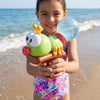 A young girl holds a green and orange ant-shaped water blaster toy with a clear sphere on a sandy beach.