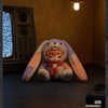 A plush doll resembling a rabbit with purple and peach ears sitting on a dark surface.