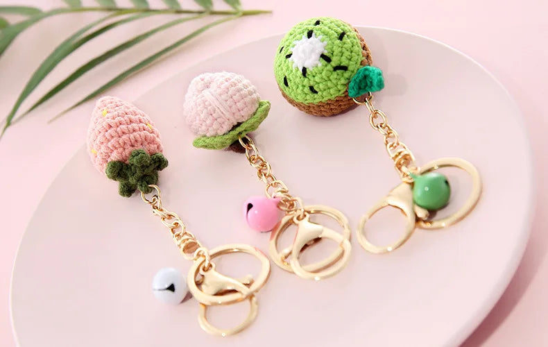 Handmade crochet fruit keychain pendants in pink and green colors displayed on a light pink plate.