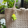 A handmade crochet dinosaur keychain in lime green and white is displayed against a beige woven bag.