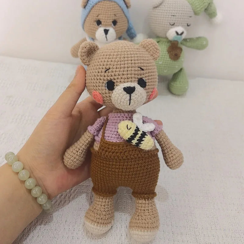 A handmade crochet stuffed bear in brown and purple clothing is shown next to two other similar plush toys.