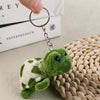 A small, green turtle plush charm attached to a metal keyring, resting on a woven surface.
