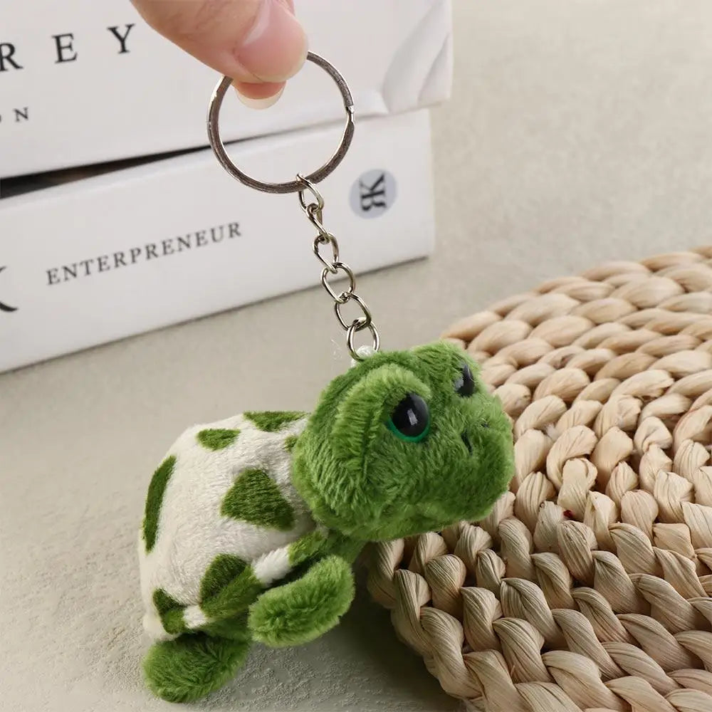 A small, green turtle plush charm attached to a metal keyring, resting on a woven surface.