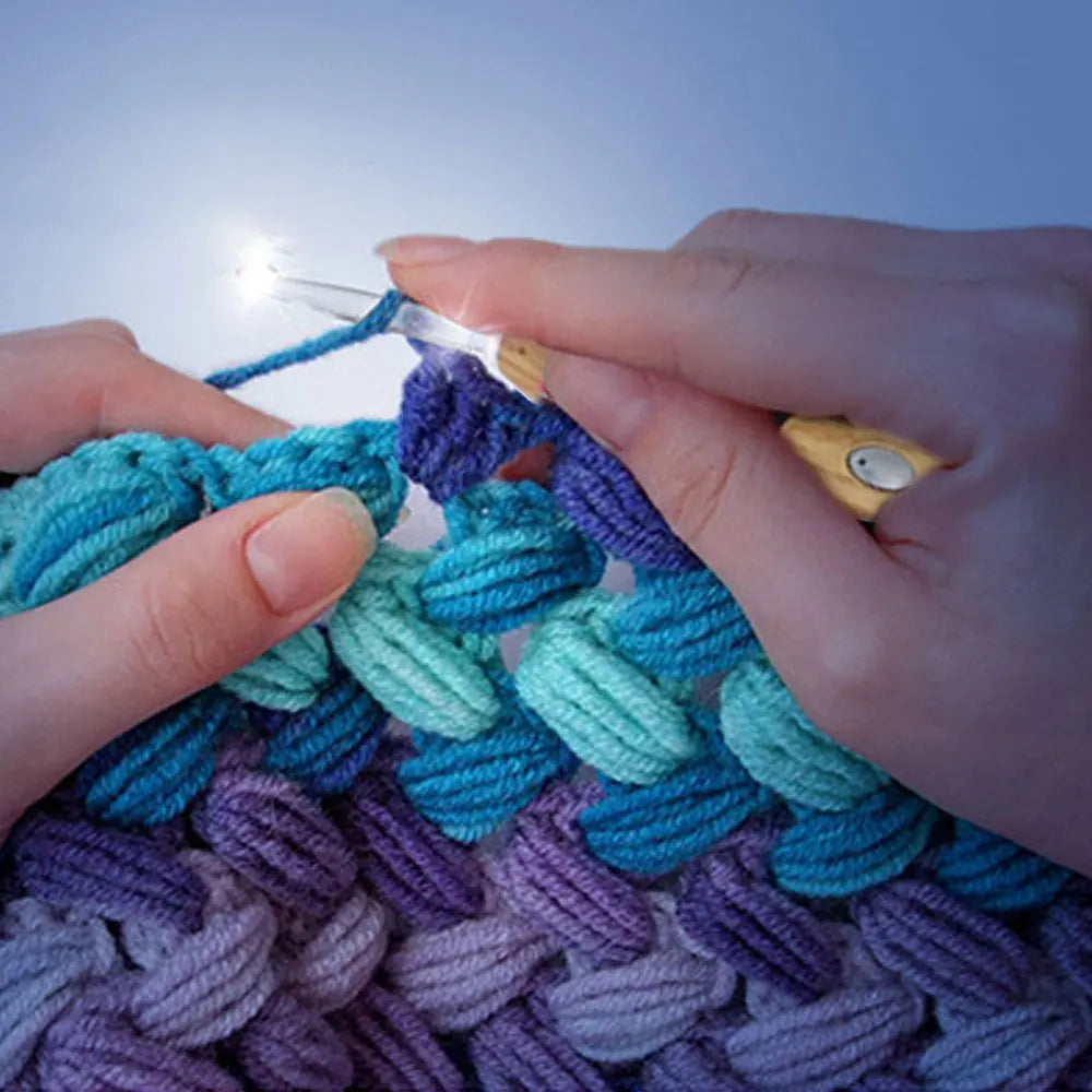 Hands are using an LED crochet hook to work on a purple and teal yarn project.