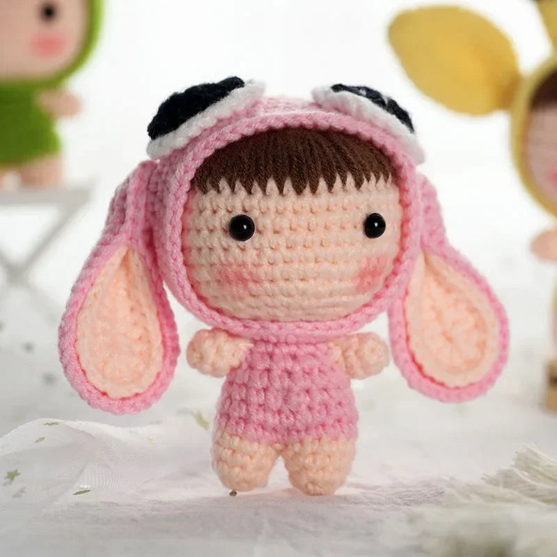 A handmade pink crochet doll wearing a pink hood with black trim, sitting on white fabric.