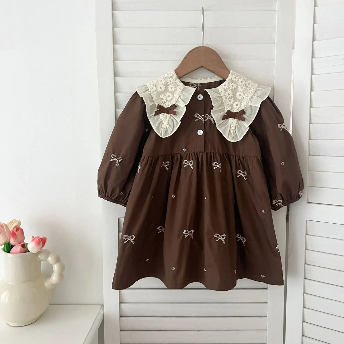 A brown cotton party dress for girls featuring lace collar and embroidered details, displayed on a wooden hanger.