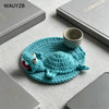 A handwoven turquoise crochet lizard coaster resting on a light gray surface next to a notebook and a small ceramic cup.