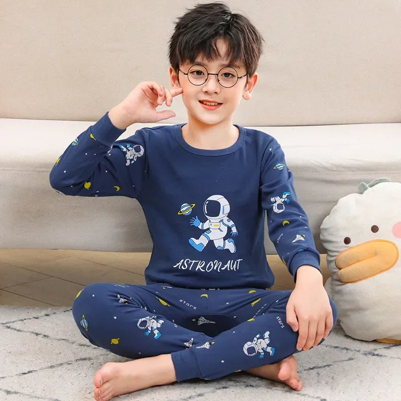 A young boy wearing a navy blue cotton pajama set with an astronaut print, sitting on the floor.