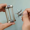 A hand holds a multi-layer stainless steel puzzle cage brain teaser with multiple vertical bars and a spherical metal object.