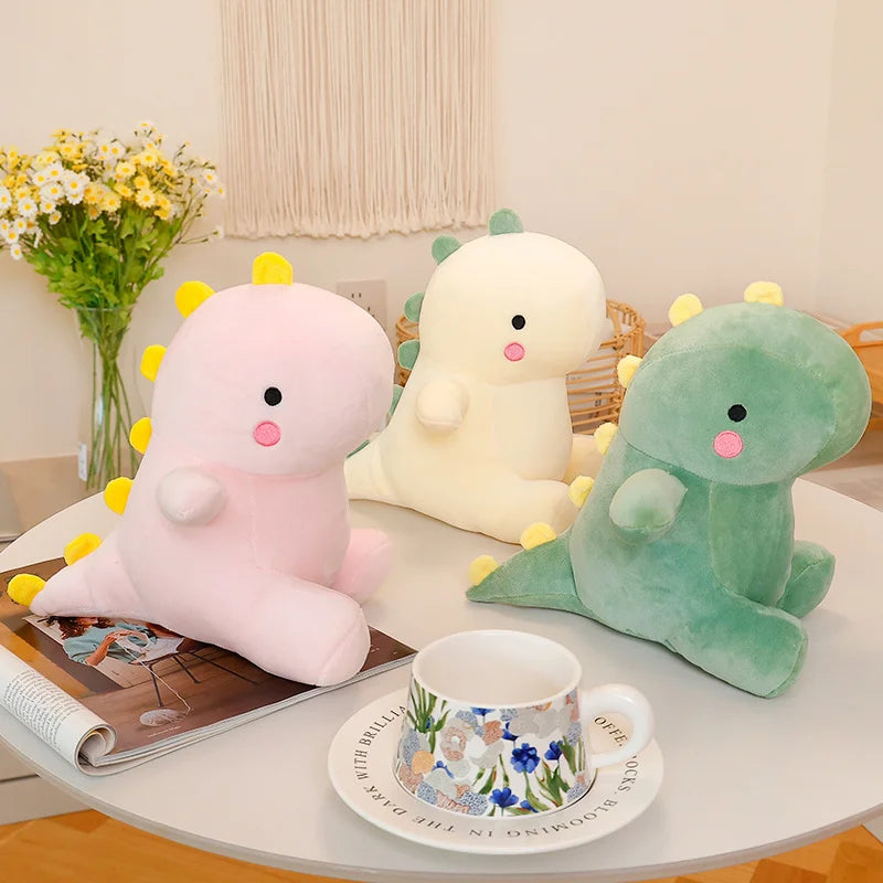 A pink and green dinosaur plush toy resting on a white table next to a mug and a book.