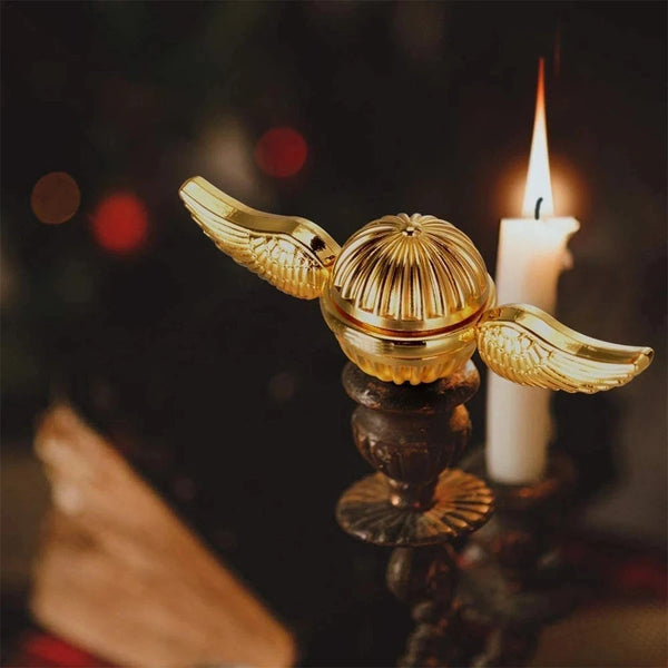 Gold angel wings fidget spinner resting on a dark surface next to a lit candle.