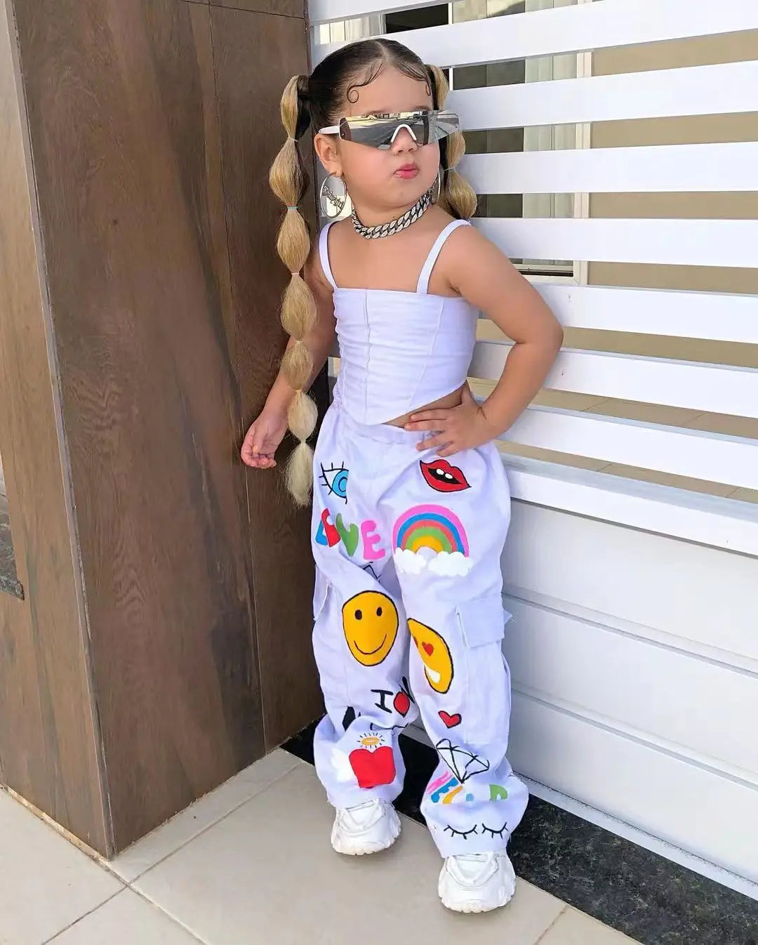 A young girl wearing a white two-piece outfit with suspender top and long pants featuring colorful graphics and sunglasses.