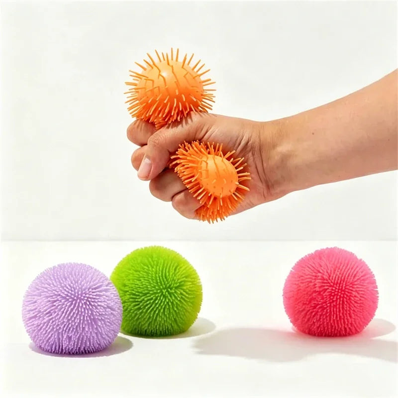 A hand holds an orange fuzz ball next to purple and green sensory balls.