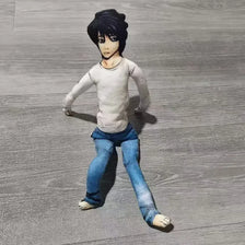 A plush doll resembling L Lawliet from Death Note, with black hair and a pale shirt, standing on a gray surface.