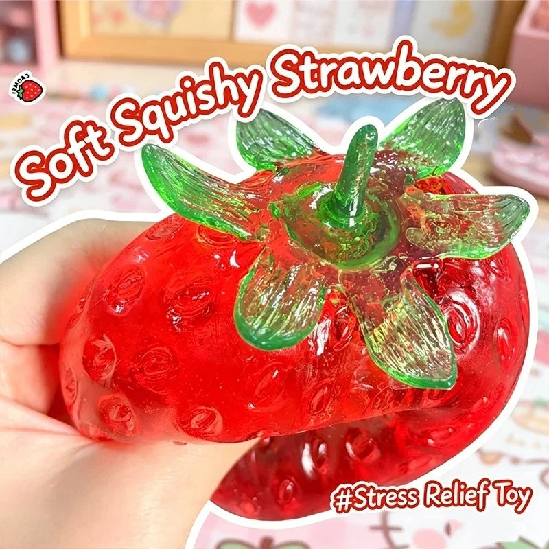 A hand holds a bright red squishy strawberry stress relief toy with green leaves.