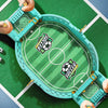 A hand holds a mini football stadium tabletop game with a green playing field and goal net on a green surface.