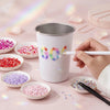 A hand holds a white tin container of colorful rhinestones next to small bowls of various colored glitter and craft supplies.