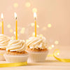 Three cupcakes decorated with white frosting are illuminated by tall yellow taper candles and bokeh lights.