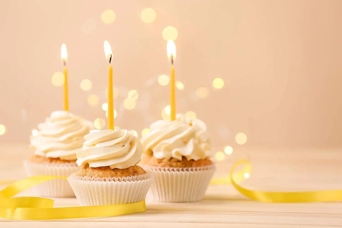 Three cupcakes decorated with white frosting are illuminated by tall yellow taper candles and bokeh lights.