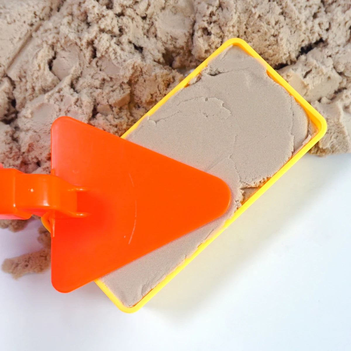 An orange plastic mold tool is shown pressing sand into a yellow rectangular mold for making sand and snow bricks.