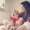 A young girl is holding a pink axolotl plush toy while sitting on a bed with an adult.