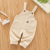 A beige baby cotton overall with adjustable straps lying on a wooden surface next to a camera.