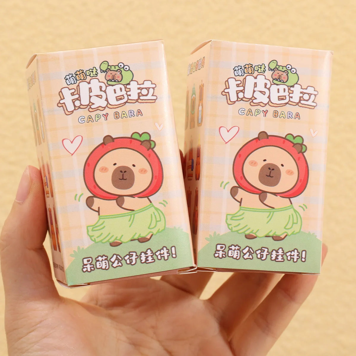يد تمسك بصندوقين كرتونيين ورديين لشارات مفاتيح (blind box) لشخصيات كابيبارا لطيفة.
