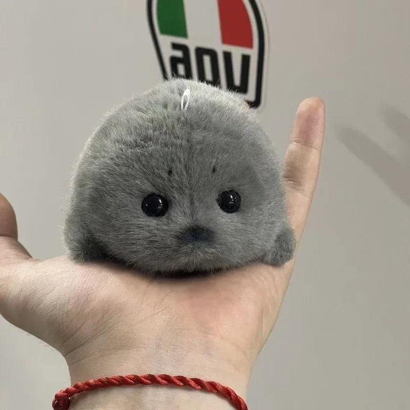A small, round grey seal plush toy is held in a person's hand against a light background.