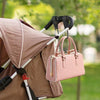 A pink leather handbag is hanging from aluminum alloy stroller hooks with nylon straps in an outdoor setting.