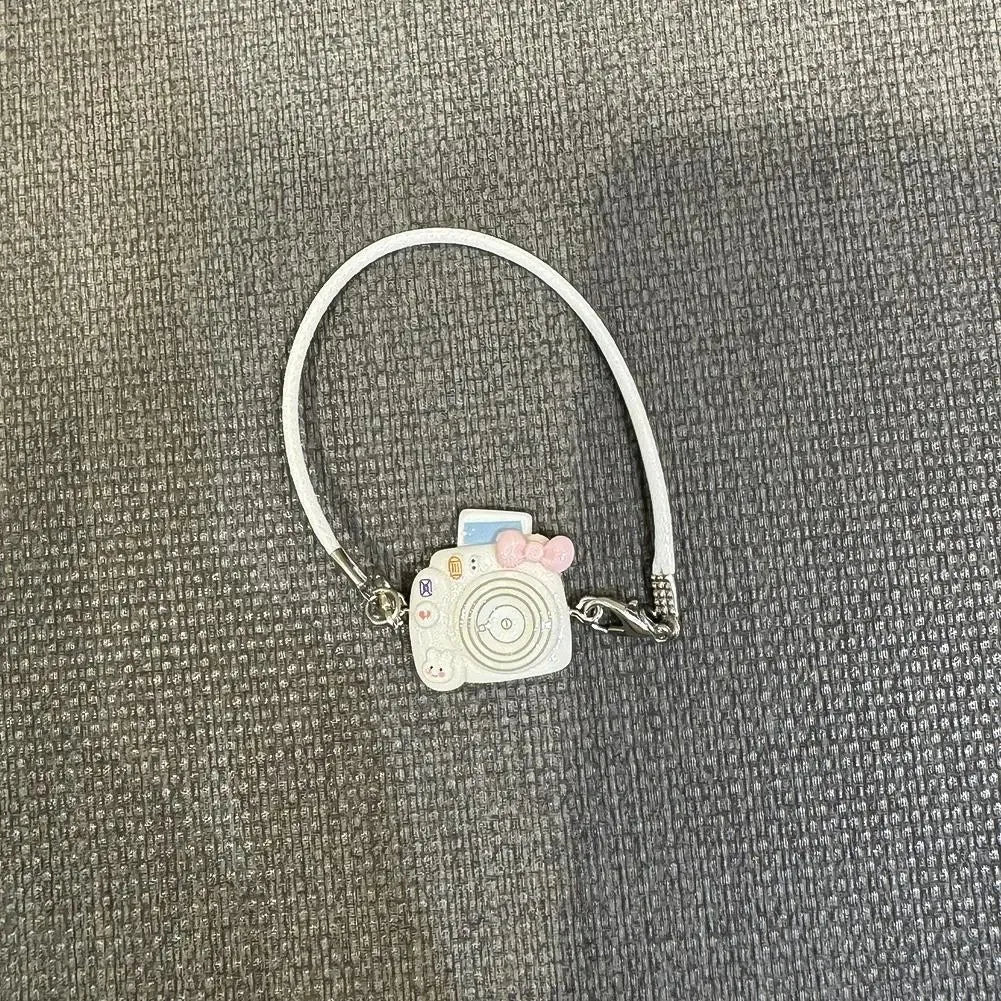 A white necklace with a small camera charm and pink accents displayed on a gray fabric background.