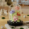A glass dome display featuring a miniature floral arrangement of pink and purple roses on a dark base.