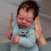A newborn baby doll with dark hair and a light blue knitted outfit is being held by a person.