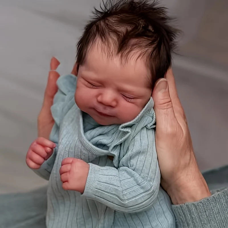 A newborn baby doll with dark hair wearing a light blue knitted outfit being held by a person.