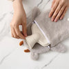 A hand is holding a light beige plush-covered hot water bottle with a zipper on a marble surface.