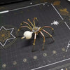 A detailed steampunk metal spider puzzle assembly kit with gold and copper colored legs and a white sphere.