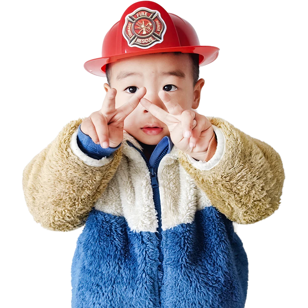 A young child wearing a red firefighter helmet and a blue and white fuzzy jacket making a peace sign.