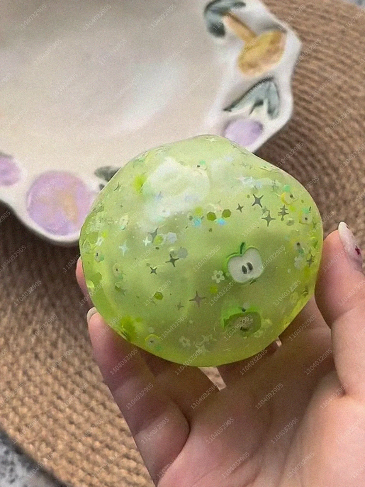 A hand holds a translucent lime green stress ball with small glittery stars and an apple design against a blurred background.