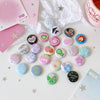 A collection of colorful kawaii mini pin badges scattered on white paper with pink and green patterned cards.