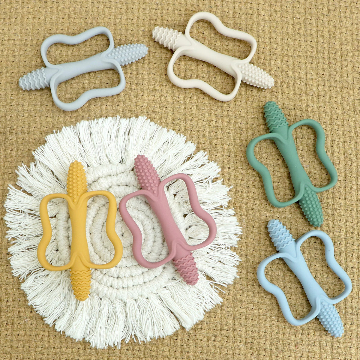 A collection of silicone teethers in various pastel colors and textures arranged on a beige fabric.