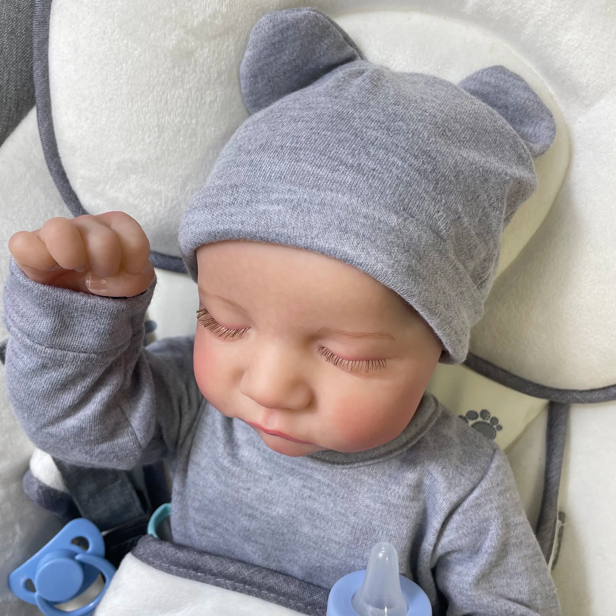 A close-up of a realistic reborn baby doll wearing a gray knitted hat and gray clothing.