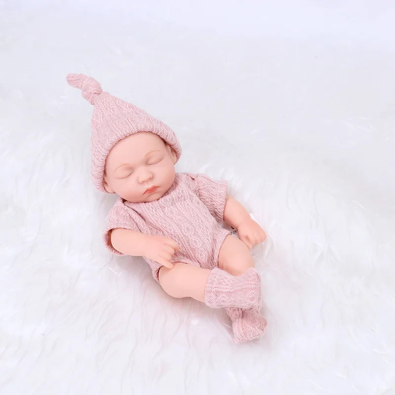 A realistic soft vinyl baby doll wearing a pink knitted outfit and matching hat, lying on a white surface.